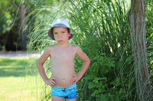 A boy in swimming trunks and a white hat stands against the backdrop of the summer sea and tropical greenery.