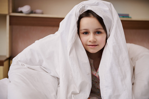 Adorable Caucasian little child girl hiding in white soft blanket, looking at camera, cutely smiling looking at camera. The concept of childhood, daily routines and bedtime
