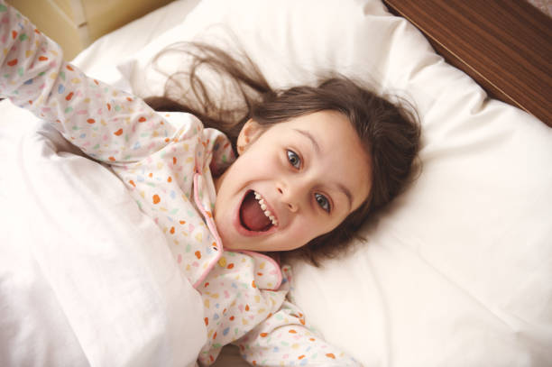 istock Mischievous little girl in pajamas, making faces at camera, stretching while waking up in morning, relaxing in her bed 1474199829