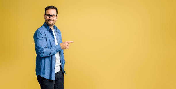 Portrait of smiling charming young adult businessman wearing denim shirt and eyeglasses pointing away at copy space isolated on yellow background Portrait of smiling charming young adult businessman wearing denim shirt and eyeglasses pointing away at copy space isolated on yellow background spokesmodel stock pictures, royalty-free photos & images
