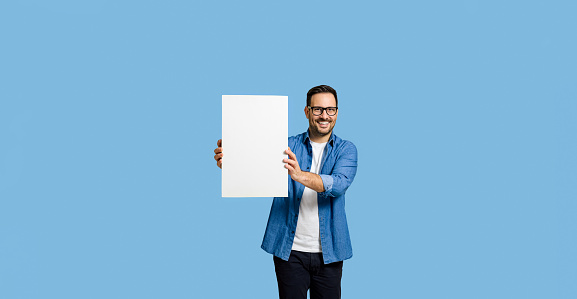 Young adult smiling businessman entrepreneur wearing denim shirt and eyeglasses showing white blank poster for marketing isolated on blue background