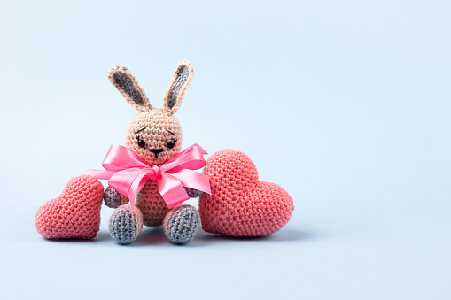 Knitted gray bunny with a crocheted pink hearts on a blue background. Knitted toys with your own hands for children or home decorations. Knitted heart is a symbol of love