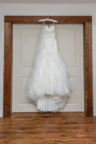 A stunning white ruffle layered style wedding gown hanging from a rustic wood bulkhead against a double doors.