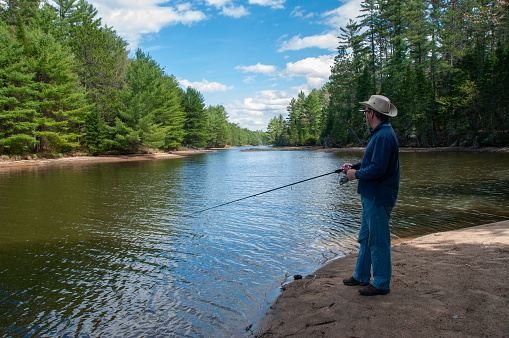 A man in a tilley hat fishes on the beach near a rural forest on the Ottawa river