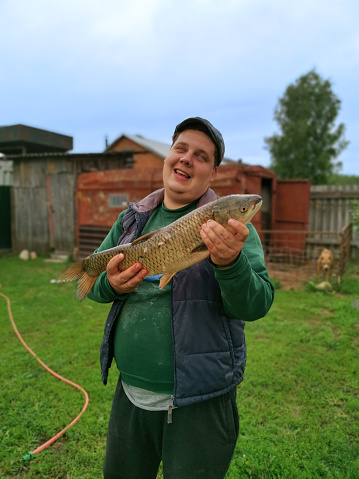 A happy male fisherman holds a big fish caught in his hands and smiles. The man caught a big fish with a line and is very happy. Hobby fishing. The man happily shows off the big fish he caught..