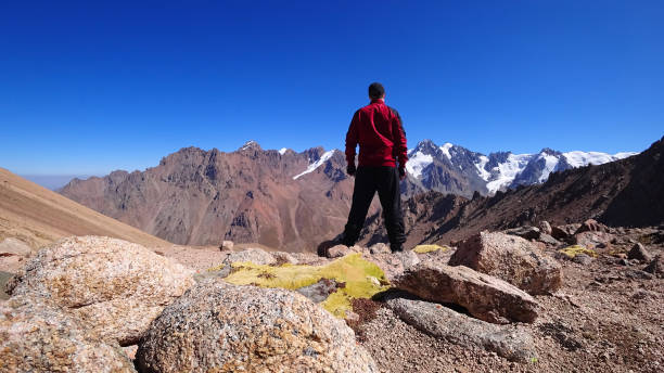 The guy looks at mountains and snow-capped peaks The guy looks at the mountains and snow-capped peaks. High mountains stretch to the blue sky. The peaks of the mountains are covered with glaciers. Steep cliffs down. There are large stones and moss mawenzi stock pictures, royalty-free photos & images