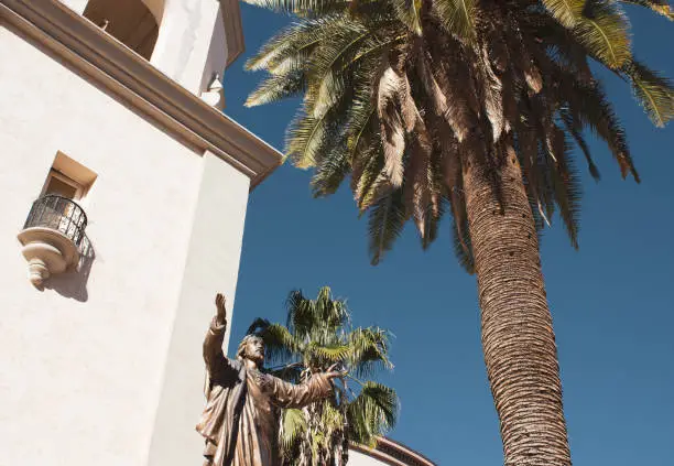 Tall Palm tree in front of a church with a statue raising it's arms up with a blue sky background