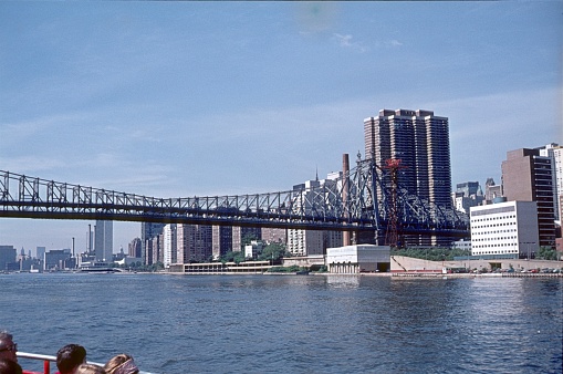 New York City, NY, USA, 1975. View of the Queensboro Bridge towards the southern tip of Manhattan from the East River. Also: Tourists on a tour boat.