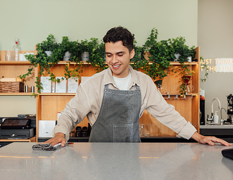 Smiling barista cleaning a counter in a cafe with a towel. Young male in an apron working as a bartender.