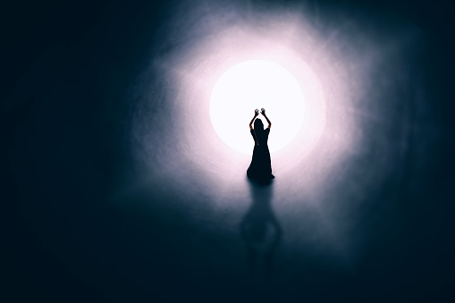 The silhouette of a woman on her knees at the light at the end of a tunnel with her arms raised in worship as she has a spiritual experience.