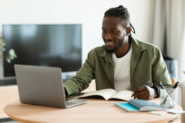 istock Excited black guy looking for job online, sitting at table, using laptop computer and taking notes, copy space 1474251106