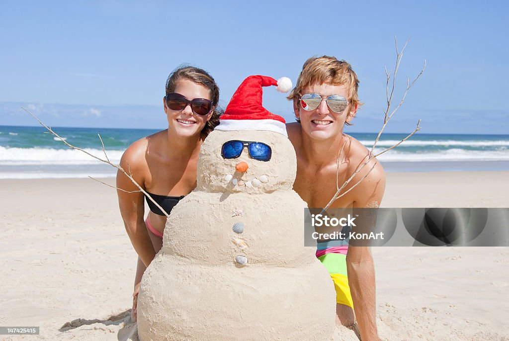 A young couple next to a sand snowman with Santa clause hat Pretty couple having fun at beach with perfectly build snowman made out of sand. With sunglasses, carrot nose and shells as buttons and mouth and santa hat. Snowman Stock Photo