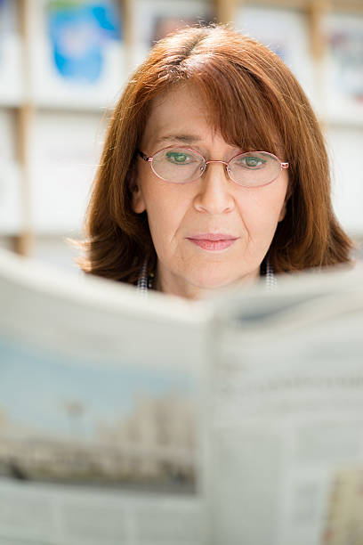istock Elderly woman with glasses reading newspaper in library 147425614