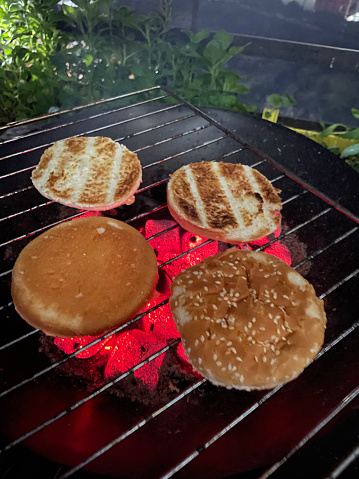 Stock photo displaying a fire pit with coals and wood that has been set alight during a dark period of the night. Burger buns with sesame seeds are being toasted over the fire. The glow of the fire and charcoal is lighting up the surroundings slightly.