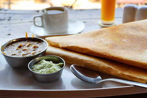Stock photo of a breakfast meal with an Indian masala dosa, a thin potato pancake that is folded and filled, it is being served on a white plate with a selection of dips / condiments / chutneys, including sambar, there is a cup of coffee in the background