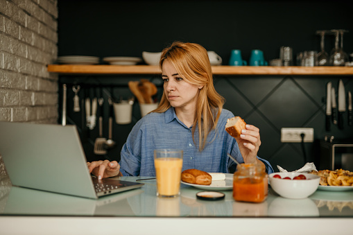 Mid-aged woman sitting alone in the kitchen, having breakfast and working on a laptop in the morning.