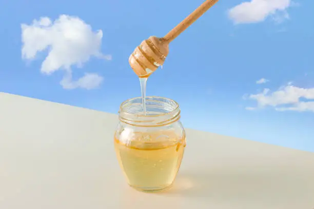Liquid golden-yellow acacia honey drips from wooden honey dipper into glass jar standing on beige table against isolated blue sky background. Concept of traditional healthy food from nature.