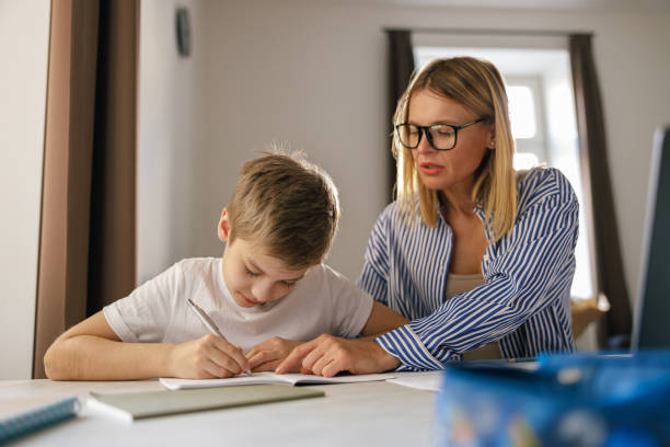 istock Schoolboy sitting at table and writing in copybook on online lesson while his mom helping him 1474308260