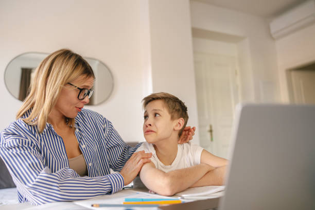 istock Tired sad kid sits at table with laptop and textbooks while mom help him with homework 1474308375