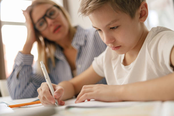 istock Schoolboy sitting at table and writing in copybook on online lesson while his mom helping him 1474310925
