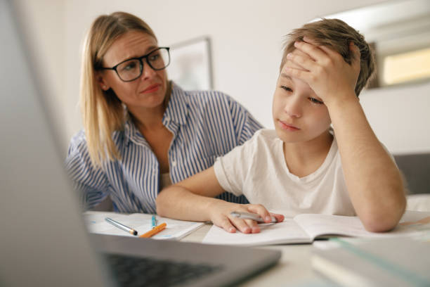 Tired sad kid sits at table with laptop and textbooks while mom help him with homework stock photo