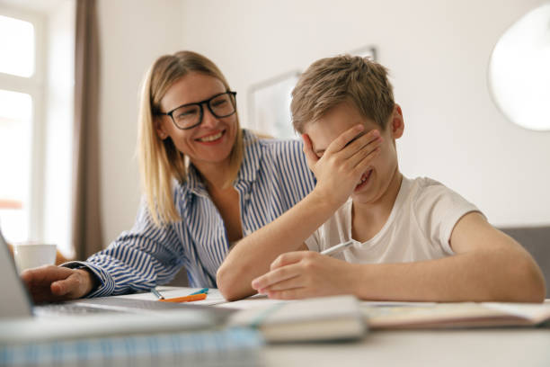 istock Tired sad kid sits at table with laptop and textbooks while mom help him with homework 1474310950
