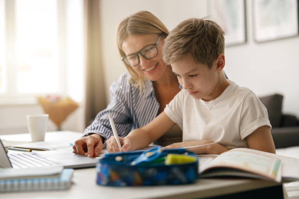 istock Schoolboy sitting at table and writing in copybook on online lesson while his mom helping him 1474310955