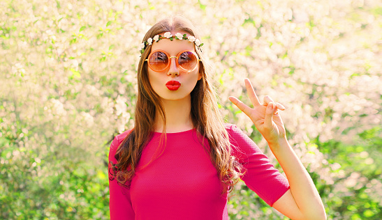 Portrait of beautiful hippie young woman blowing her lips sending air kiss wearing floral headband, sunglasses in spring blooming garden on flowers background