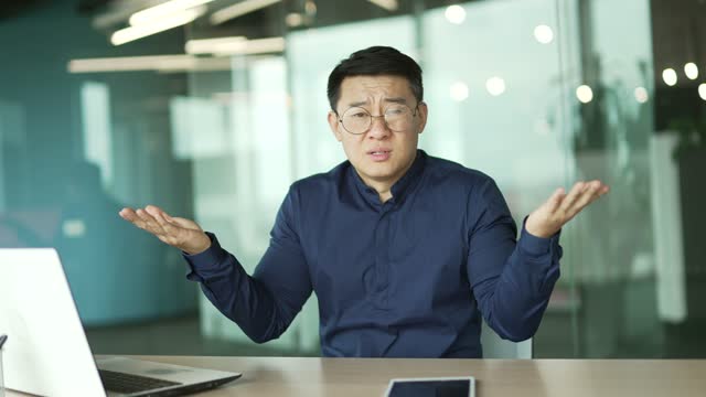 Puzzled asian businessman employee sitting next to laptop computer and shrugging shoulders in hesitation doubting incertitude gesture uncertain reaction while looking to camera