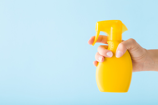Young adult woman hand holding yellow spray bottle of sunscreen or cleaning detergent on light blue wall background. Pastel color. Closeup. Empty place for text. Side view.