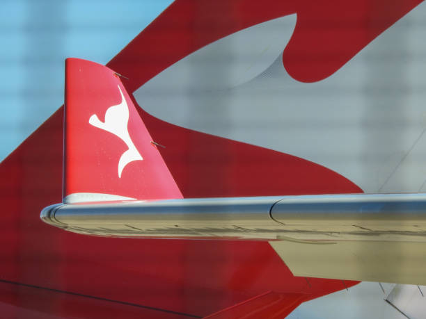 Qantas logos on planes parked at the maintenance area near the domestic terminal of Sydney Kingsford-Smith Airport.  The closer object is the wing tip of an Airbus A330-203, registration VH-EBL.  The far object is the vertical stabiliser of an Airbus A380-842, registration VH-OQL.  The checked pattern lines are the security fence.  This image was taken near Joyce Drive, Mascot on a sunny and hot afternoon on 18 March 2023.