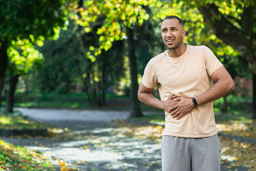 Latin american runner sportsman has severe stomach pain, man holding hand on side of stomach after doing exercise and fitness in park outside.