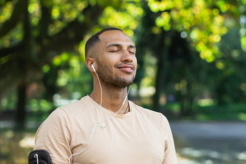 Close-up portrait of sportsman in park, hispanic man jogging in park with eyes closed breathing fresh air and resting, jogging with headphones listening to music and online radio and podcasts.