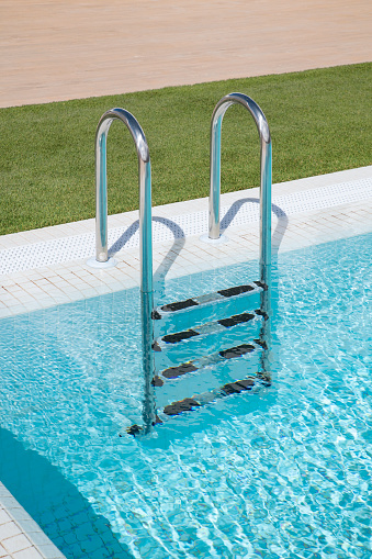 Sturdy ladder in a sleek design and with non-slip rungs to ensure safety and ease to use, positioned at the edge of the crystal clear swimming blue pool, convenient entry and exit point from the water.