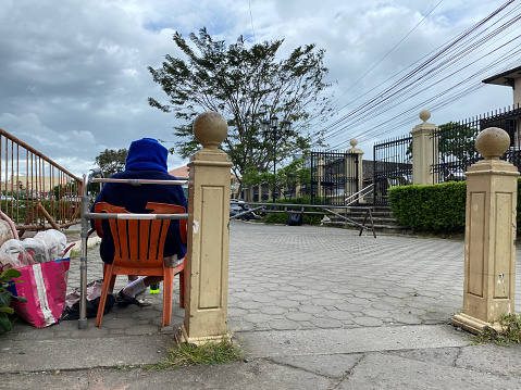San Pablo City, Laguna, Philippines - February 25, 2023: Old female beggar wearing a blue jacket seated in a wheelchair on the churchyard pavement  POV