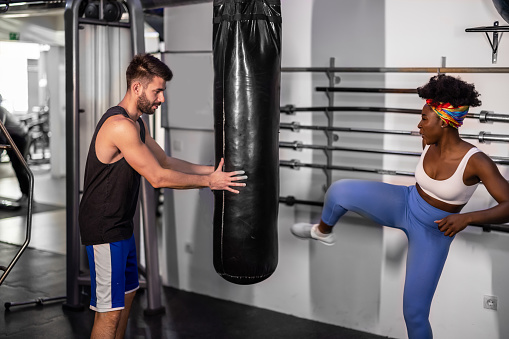 A young training coach is assisting a female African athlete while she is punching a bag.