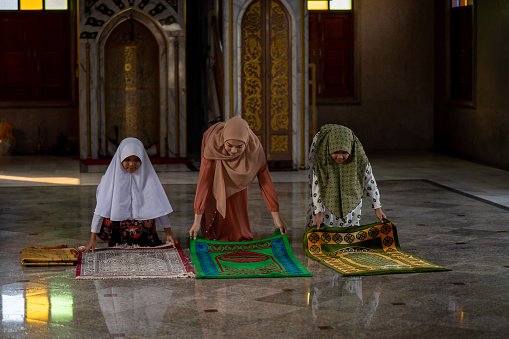 A beautiful Muslim girl in a hijab teaches children how to lay carpets to pray in the mosque.