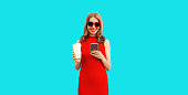 Portrait of beautiful smiling woman with smartphone with cup of coffee wearing red dress and heart shaped sunglasses on blue background