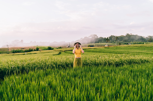 Cheerful young lady in summer clothes enjoying lush exotic nature while taking stroll about green rice fields in Bali countryside during summer vacation