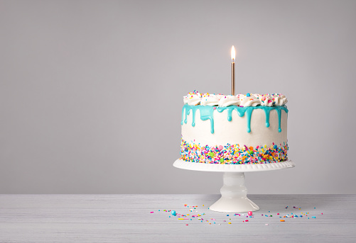 Vanilla buttercream birthday cake with a teal blue ganache drip, lit gold candle, and colorful sprinkles on a light grey white background. Copy space.