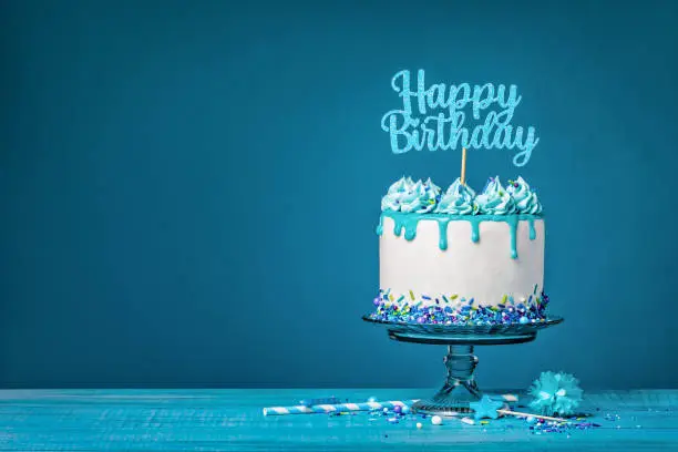 White drip cake with teal ganache, sprinkles, and a happy birthday topper over a blue background. Simple and trendy. Copy space.