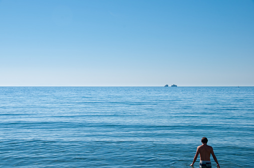 A young man stands about to swim in the blue waters of the Mediterranean Sea