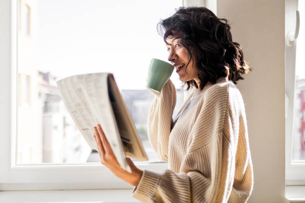 mujer joven bebiendo una taza de café leyendo el periódico - mujer leyendo periodico fotografías e imágenes de stock