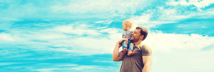 Happy strong father holding his son child on shoulder outdoors on blue sky background with clouds, blank copy space for advertising text