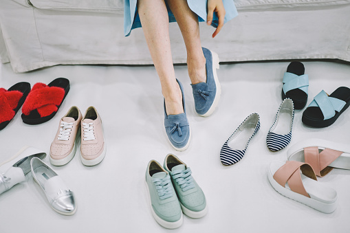 Woman trying on blue denim moccasins choosing from different pairs of colorful trendy and casual types of footwear beside on floor