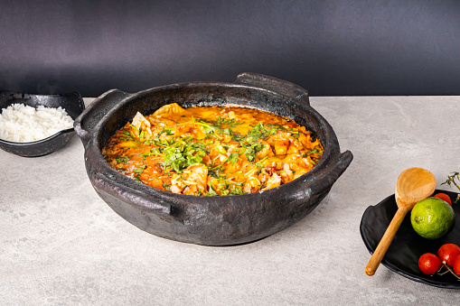 Delicious Brazilian fish moqueca with tomato, onion, olive oil, coriander and orocum seed. Made in clay pot on white table and black background. With rice.