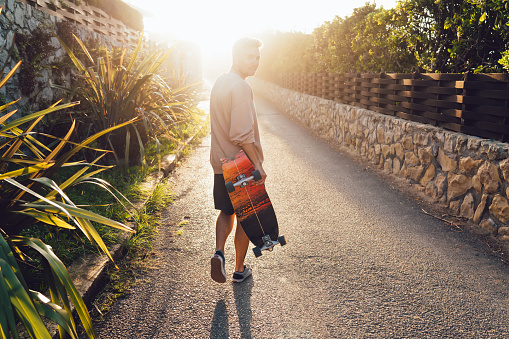 Back view of anonymous male with longboard in casual wear strolling along narrow pathway among green plants and looking over shoulder