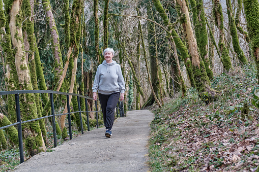Senior active caucasian woman with gray hair cheerfully walking along a tourist trail in a relic forest. Full-length front view. Yew-boxwood grove, Caucasian Nature Reserve, Russia.