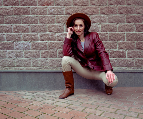 A young woman in a leather jacket sits near a gray brick wall. Portrait of a pensive woman in a hat and brown boots. Brick wall background. Woman crouched in light pants