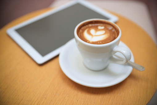 White cup of Hot coffee beside digital tablet  on wooden desk. Simple workspace or coffee break in morning/ selective focus